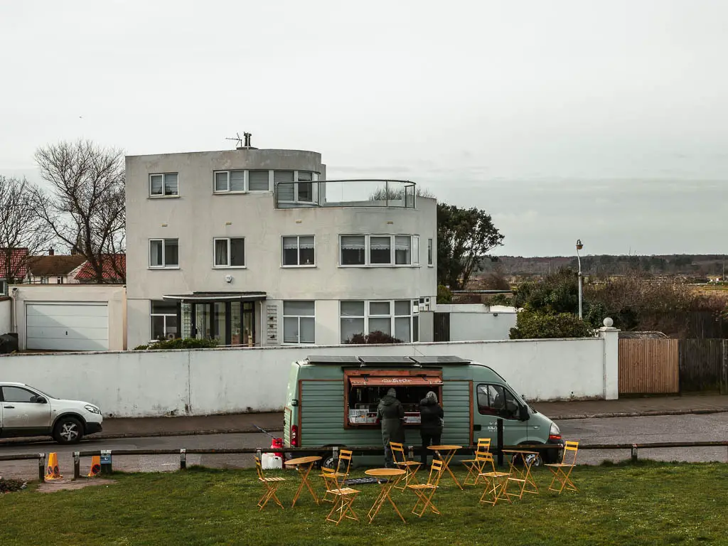 A coffee truck next to the green on the walk towards Sandwich from Deal. There are yellow tables and chairs on the green. There are two people standing at the coffee truck. 