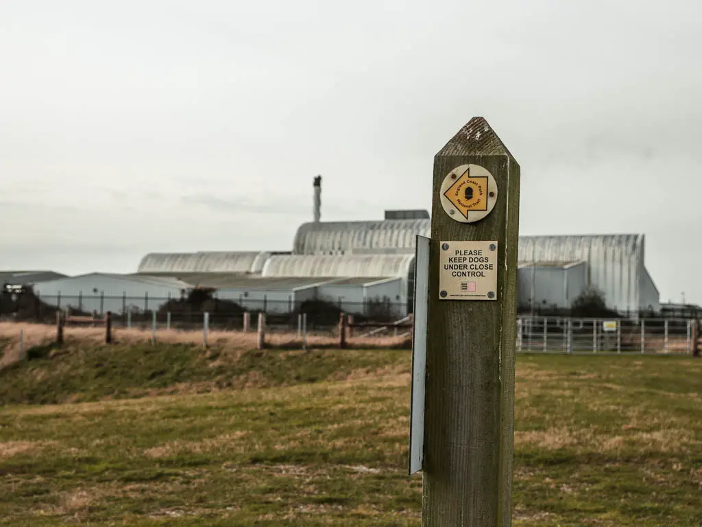 A wooden stump signpost with a yellow arrow. There is a large industrial building behind it. 