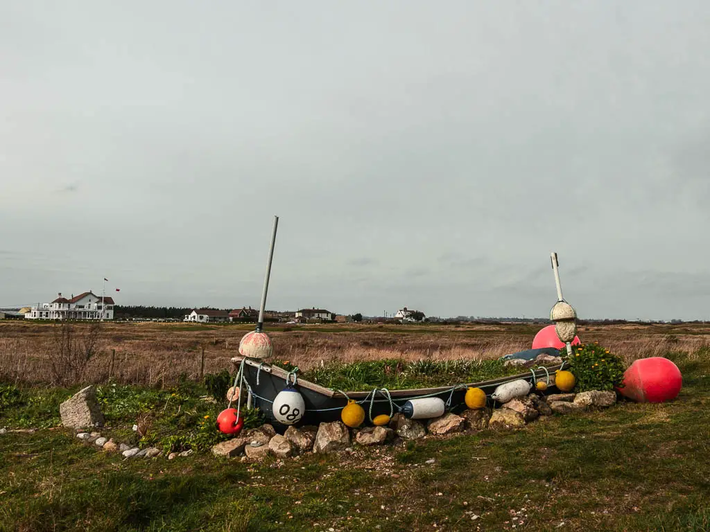 A black canoe decorated with colourful buoys on the walk from Deal to Sandwich. It is in front of a large field with some white coloured buildings on the other side. 