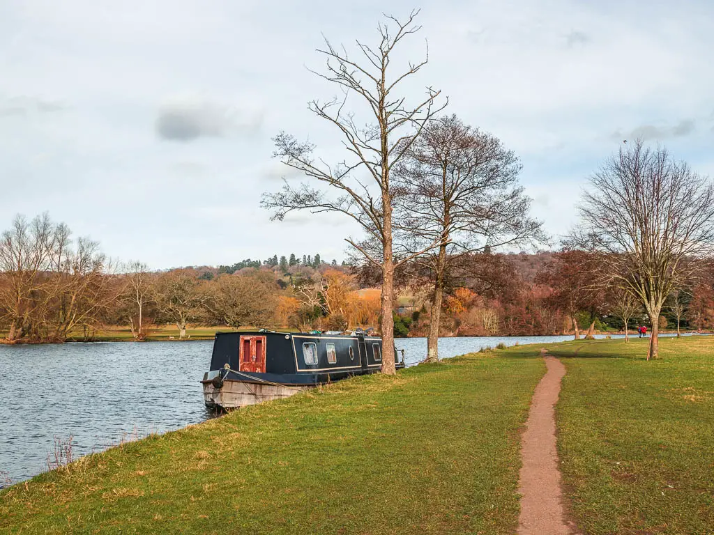 A narrow trail through the green next to the river. There is a barge moored on the side of the river. 