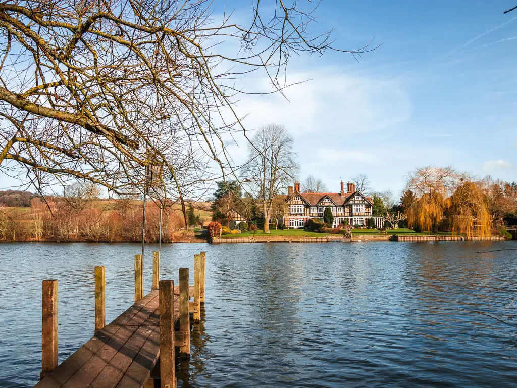 Looking across the River Thames to a large manor house on the green on the other side.  There is a wooden plank leading into the river. 