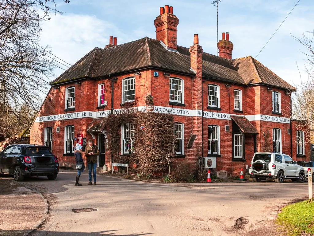 The corner of a large red bricked pub in Aston. There are a couple of cars parked outside on the road and a couple of people standing talking on the road. 