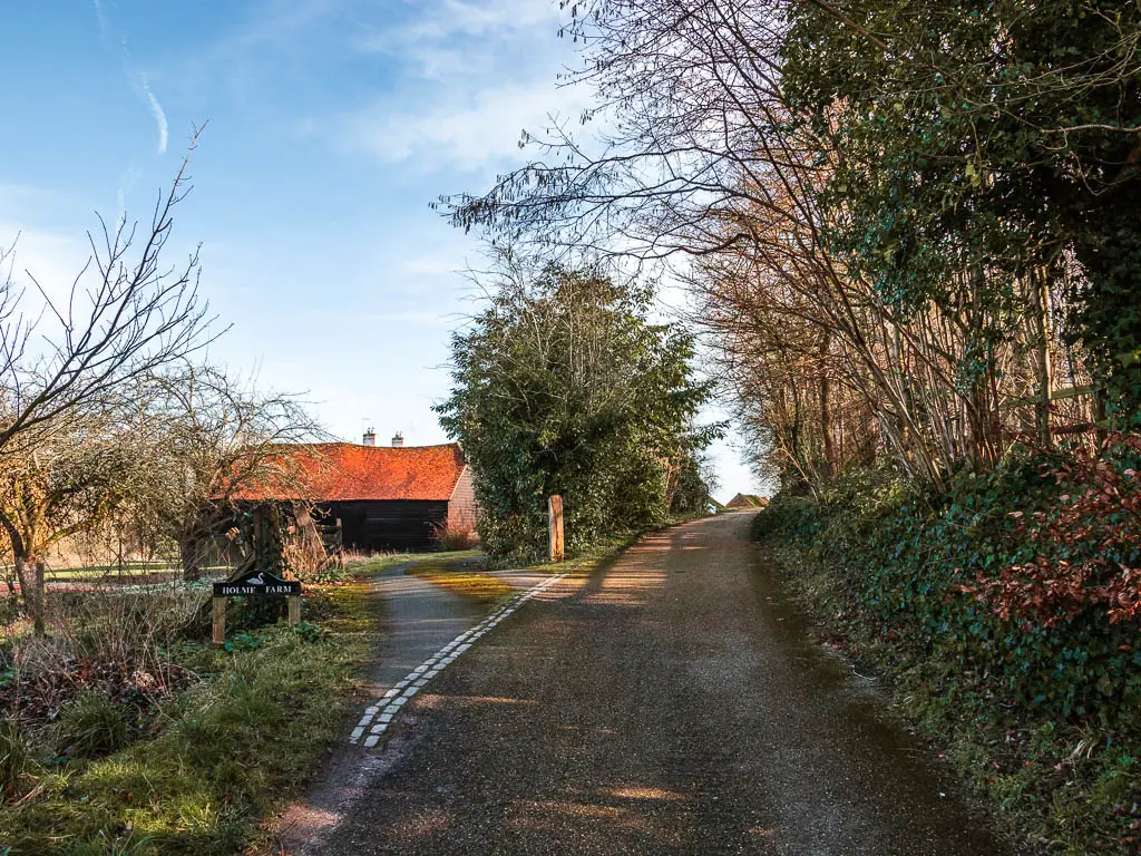 A back road leading uphill, with as driveway branching off there left and leading to a red roofed building. 