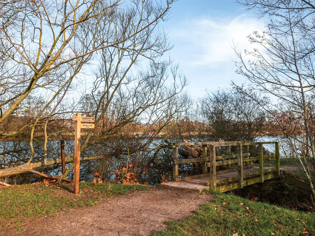 A dirt trail leading over a wooden bridge next to the River Thames on the walk between Henley and Marlow.  There is a wooden signpost next to the trail. 