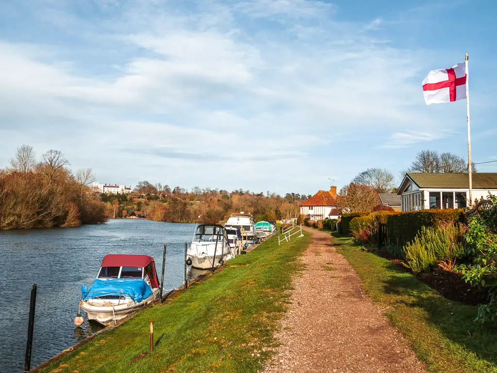 A gravel dirt trail with a sloping grass bank to the left and onto the river. There are a few boast moored to the bank. There are a couple of houses on the right with the St George's flag flying. 