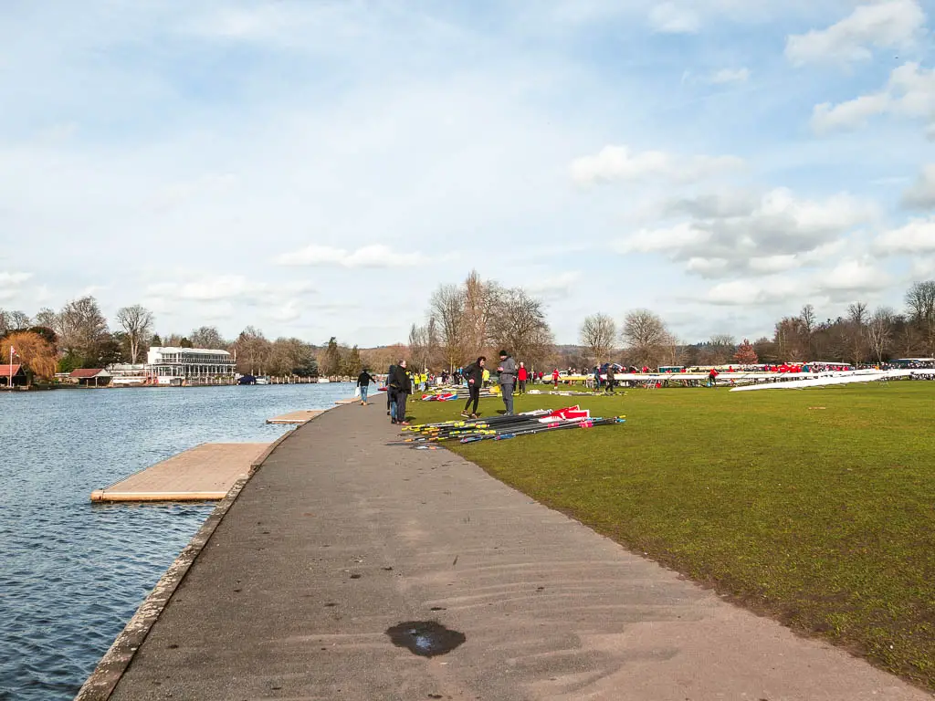 Looking along the asphalt path with the river on the left and green on the right with people preparing their row boats.