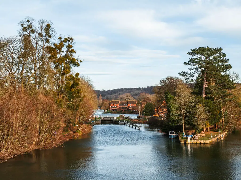 Looking down along the River Thames on the walk from Henley to Marlow.  The river is lined with trees, and ahead is a lock and some red house facades in the distance.