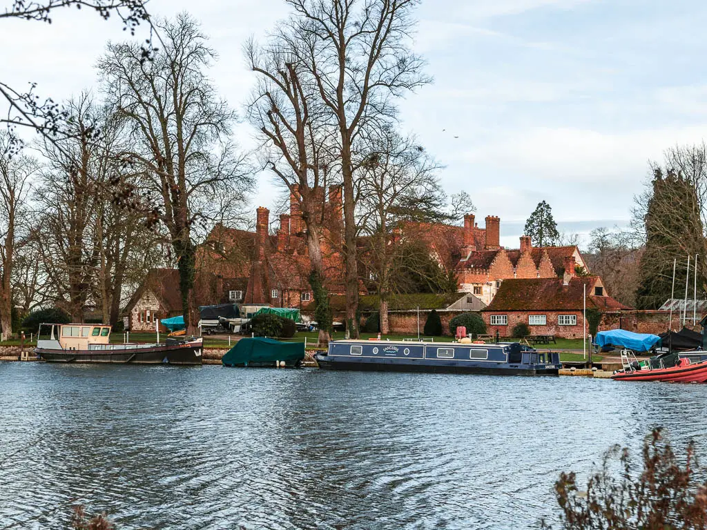 Looking across the river to a large Manor House with a light red facade and boats and a barge moored along the bank.