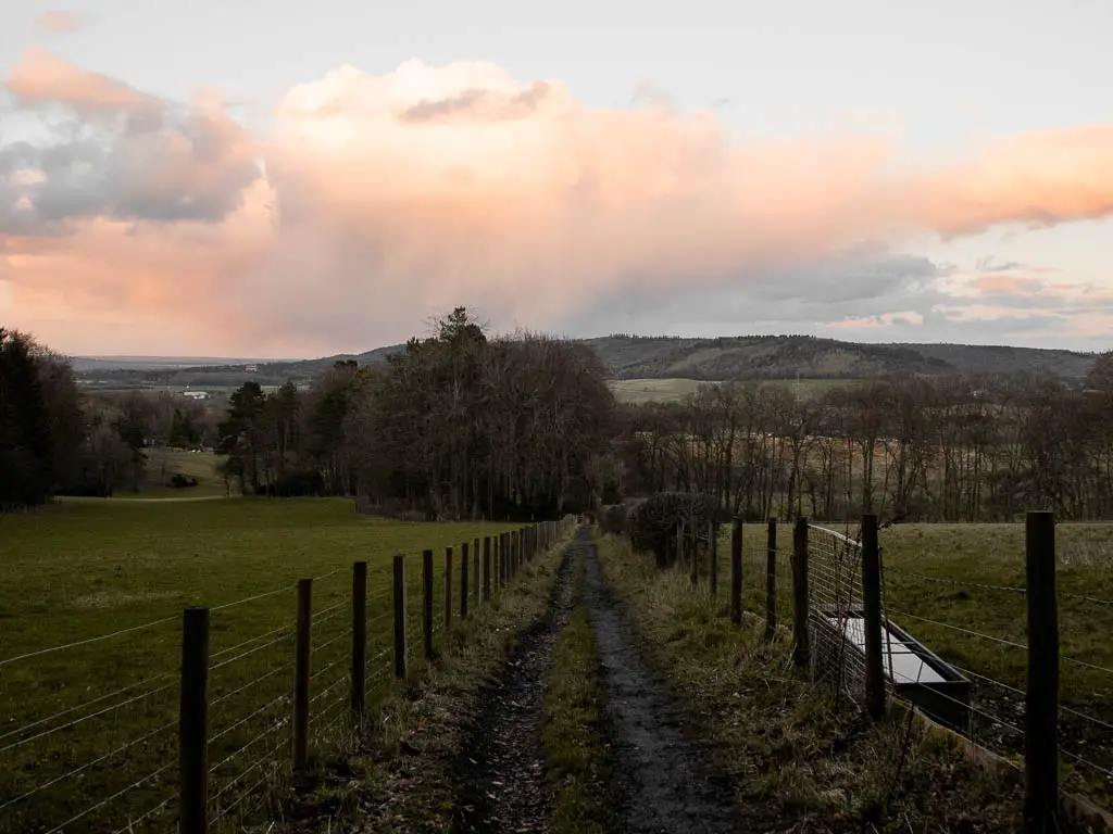 A muddy trail leading downhill towards some trees and a view of the hills in the distance. The trail is lines with a wooden and barbed wire fence. 