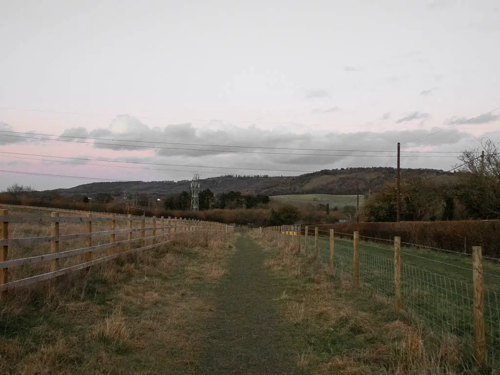 A grass trail with a wooden fence on the left and barbed wire fence on the right. There are hills in the distance. 