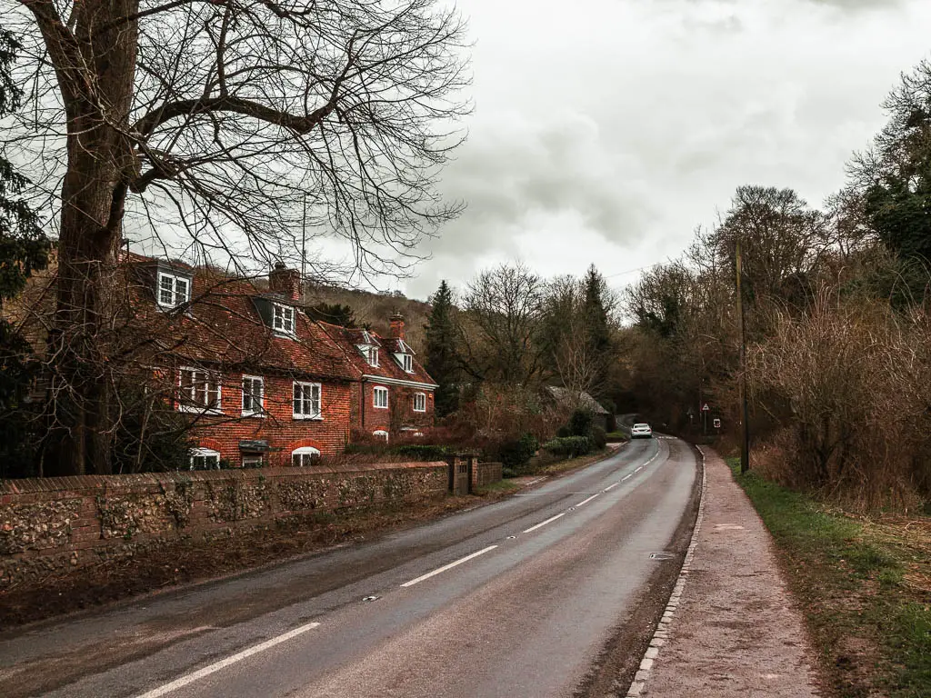 A road with a red bricked house on the left side of it. There are a few trees next to the house and lining the right side of the road.