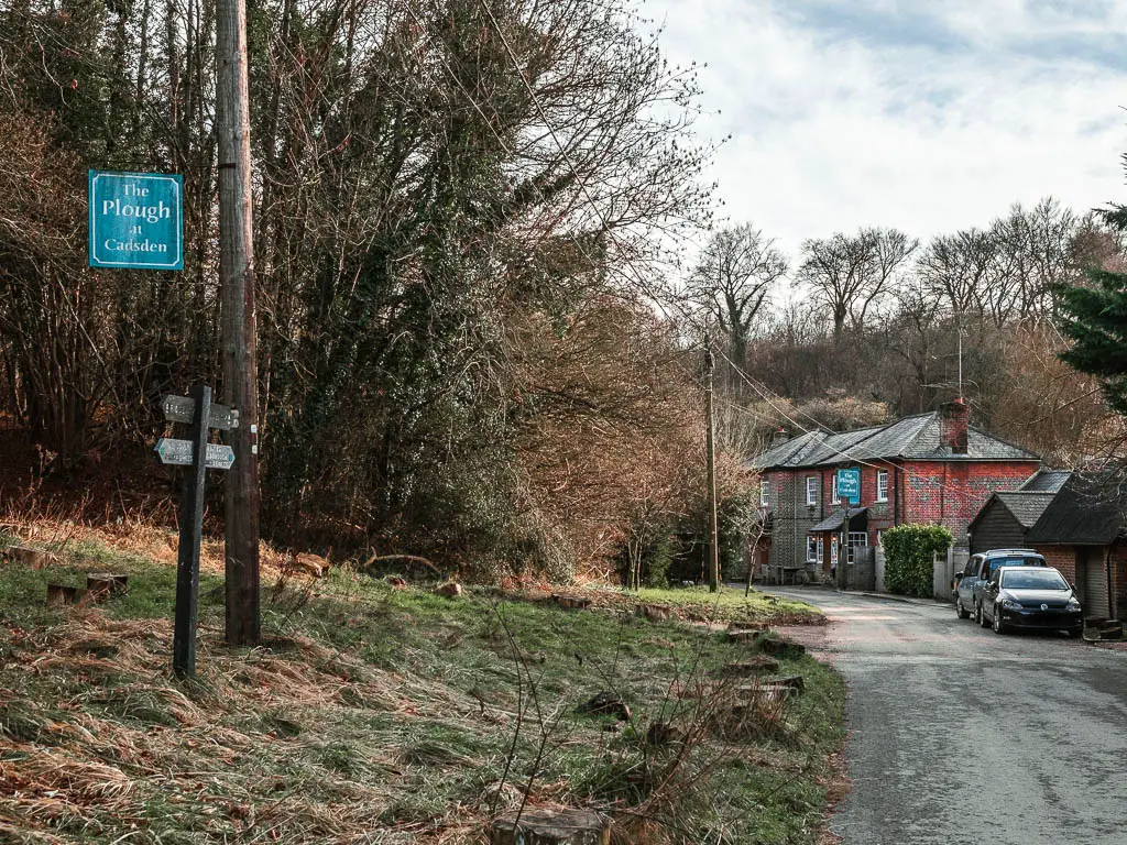 A road leading to a red bricked pub. There is a grass bank on the left side of the road with a wooden trail signpost. 