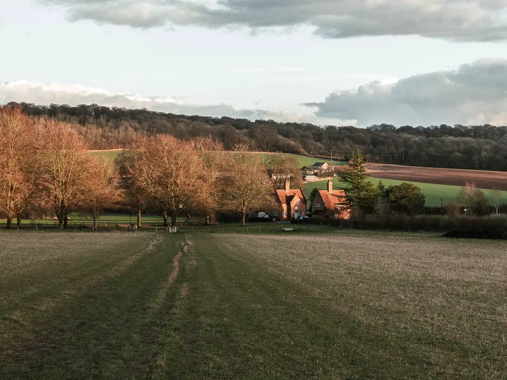 A large field leading downhill towards two red bricked houses and some trees on the walk back to Wendover from Coombe Hill, Beacon Hill and Whiteleaf Hill. 