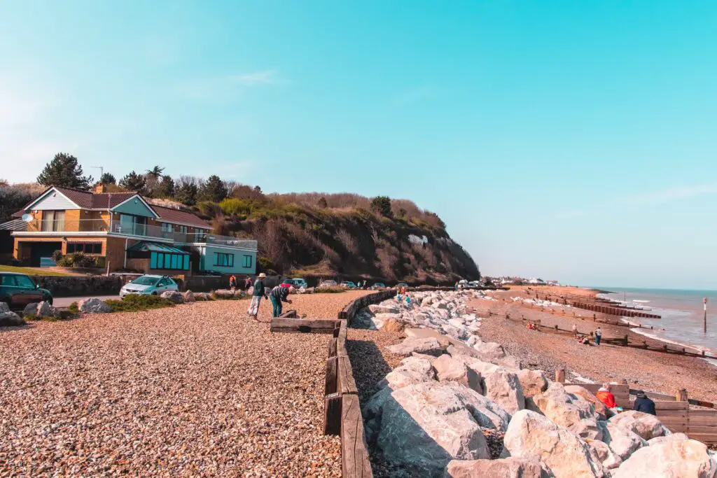 A shingle beach on the England Coast path when walking from Dover to Deal. There are a few people on the beach, the sky is blue and there is a tree bush covered cliff up ahead.