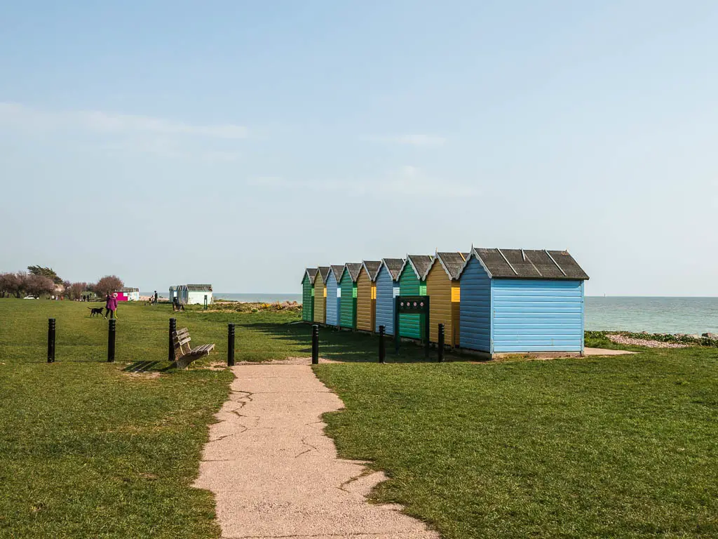 A path through the green with blue, yellow and green beach huts facing the sea.