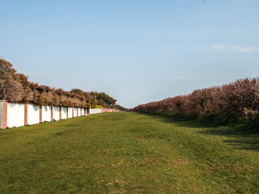 A large long stretch of green with a white wall to the left and bushes to the right. 