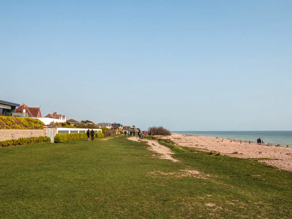 A large green next to the shingle beach. There are lots of people walking ahead on the green. 