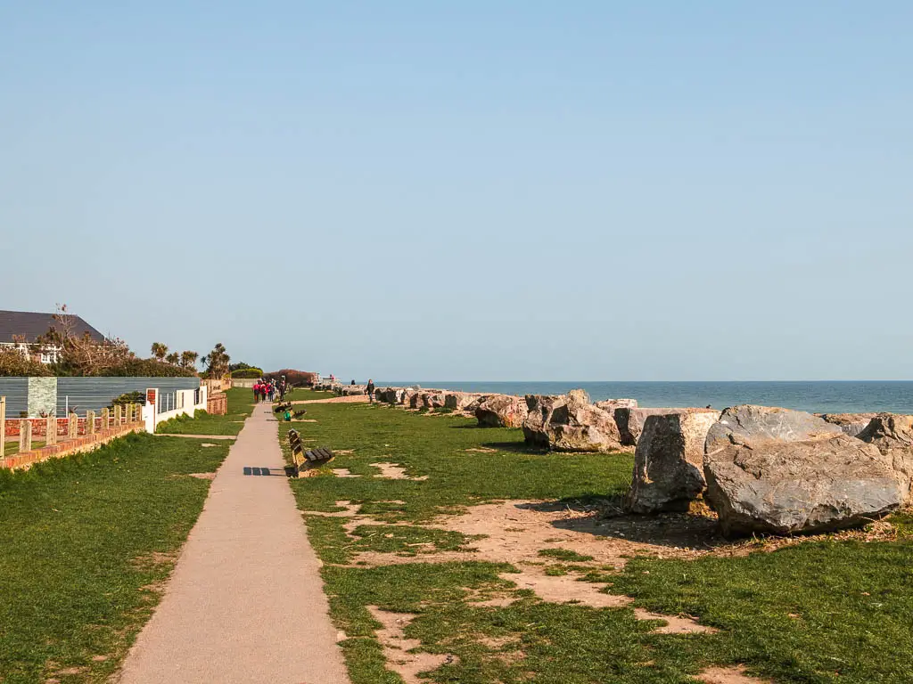 A straight trail on the left with green on the right and a neat row of large boulders. There are a few benches lined along the green. 