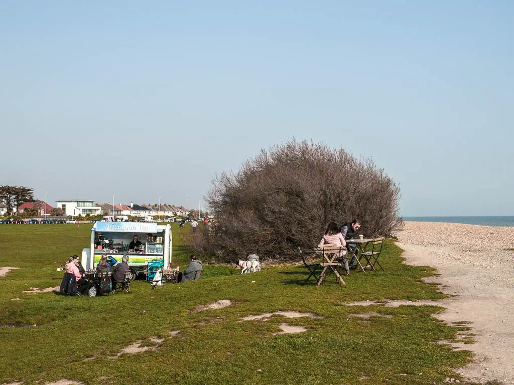 A small food truck on the green on the left and the beach to the right. There is a table and bench on the green with a couple sitting. 