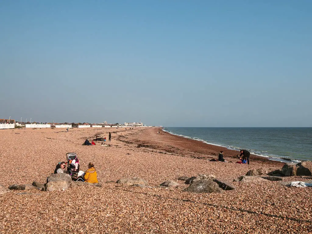 A long stretch of shingle beach with the blue sea to the right. there are a few people sitting on the beach.