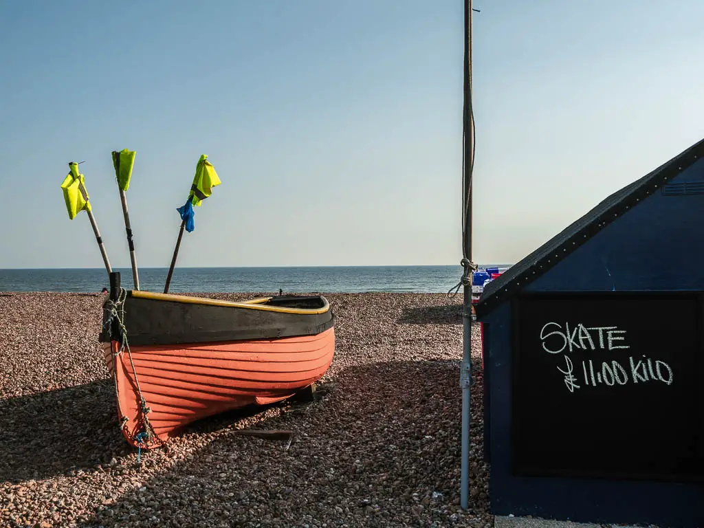 A red boat on the shingle beach with yellow flags on the walk from Littlehampton to Worthing.