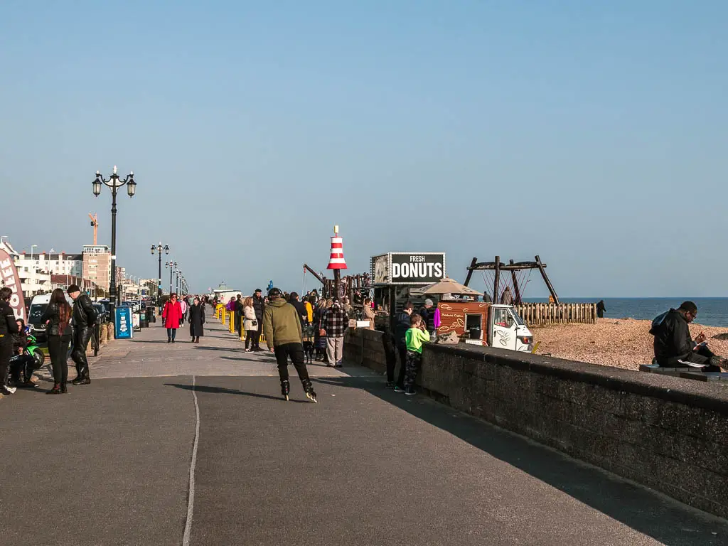 The concrete promenade with lots of people walking on it in Worthing. There is a donut stall on the right whit a black sign.