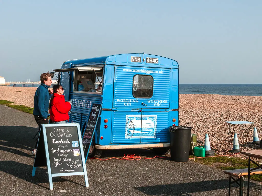 A blue gin truck next to the shingle beach in Worthing. There are a couple of people ordering from the truck. 