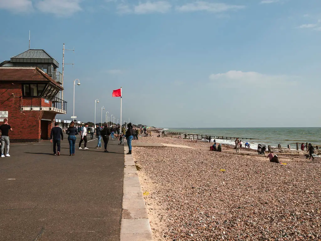 the promenade on the left and shingle beach on the right on the walk through Littlehampton. there are lots of people walking along the promenade. 