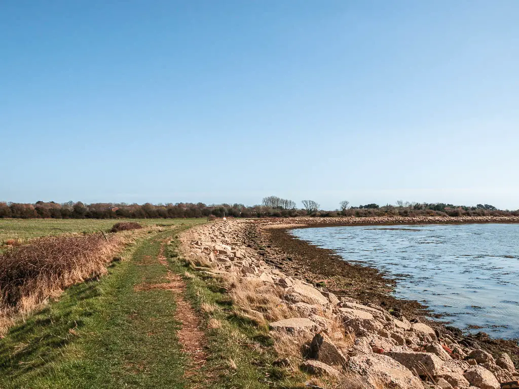 The grassy coast path on the left with rocks to the right and then the blue sea when walking around Thorney Island.