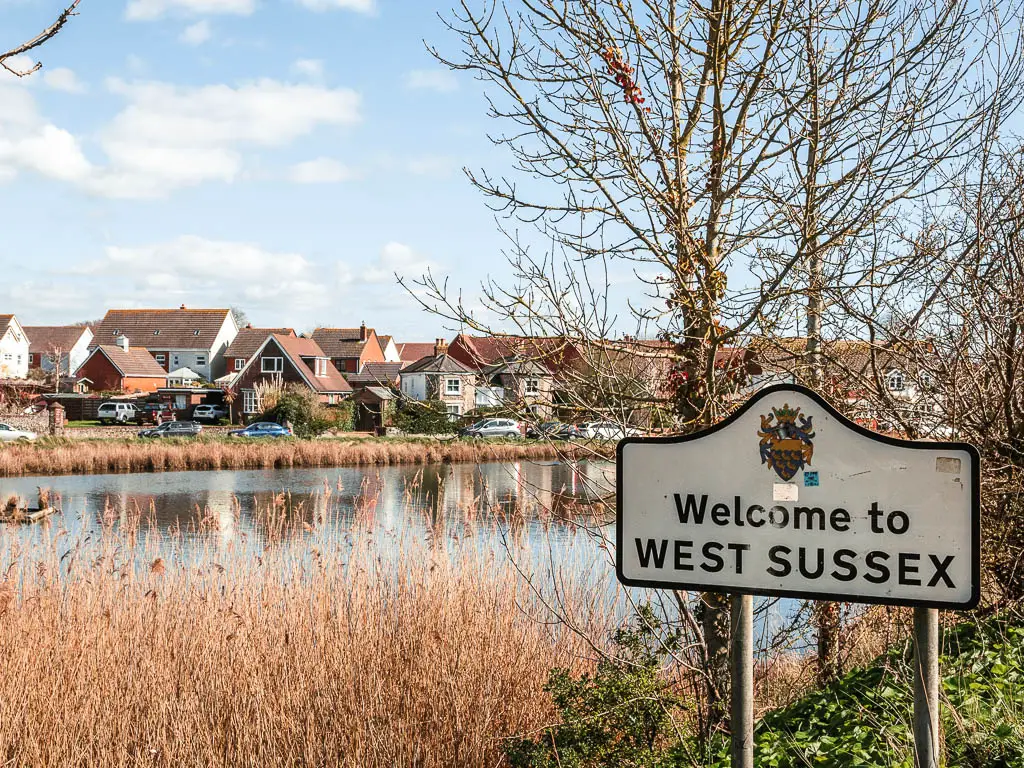 A 'welcome to West Sussex' sign in front of the lake on the walk to Thorney Island. There are houses on the other side of the lake. 