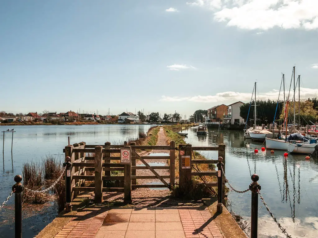 A wooden gate leading to a trail across the lake. There are sail boats moored to the right.