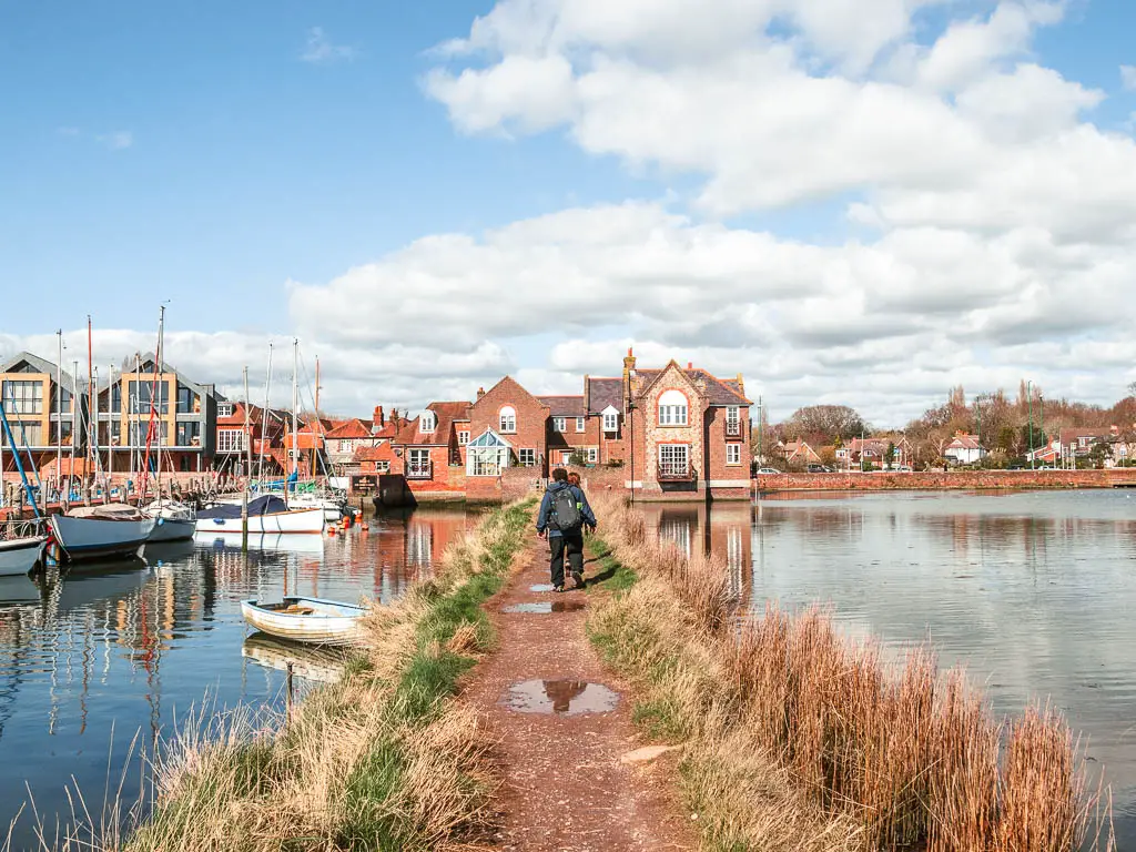 Two people walking along the path over the lake. There is a building at the end and sail boats moored to the left. 