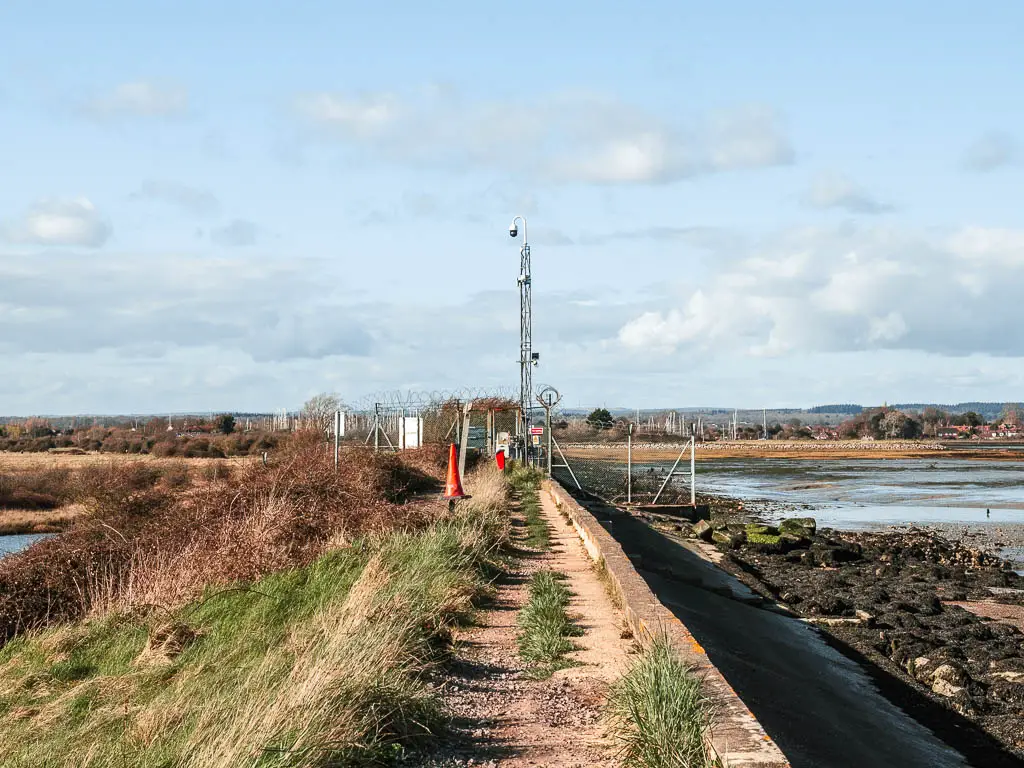 A narrow gravel type path leading towards a metal fence and gate. There is overgrown grass to the left of the path and the sea wall to the right.