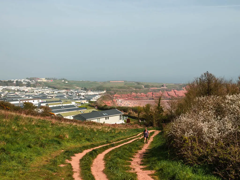 The trail leading downhill through the grass with a holiday park ahead, and red cliffs just visible in the distance. 