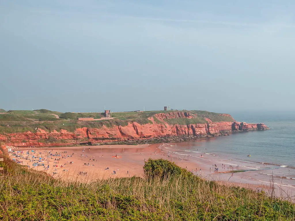 Looking across the green to the red sand beach below and red cliffs on the other side on the walk from Exmouth to Budleigh Salterton along the coast path. There are lots of people down on the beach.