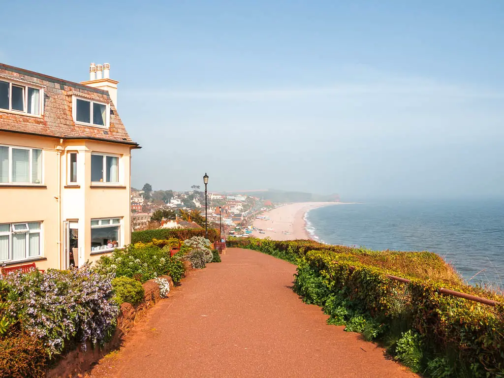 A wide path with cream walled building on the left Budleigh Salterton Beach stretching ahead at the end of the walk from Exmouth. 