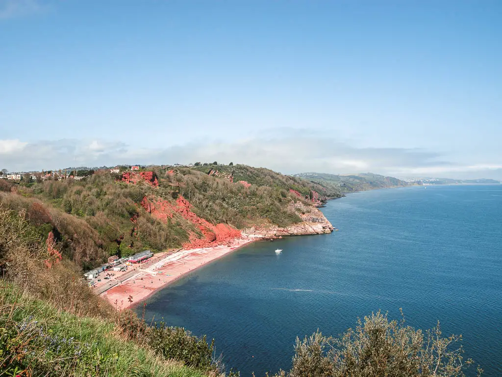 Looking down to the blue sea where it meets the beach which is surrounded by a tree covered cliff in Babbacombe, at the start of the walk towards Shaldon and Teignmouth. 