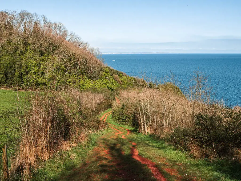 A red coloured dirt trail snaking down the grass hill, surrounded by bushes with the blue sea ahead on the coastal walk from Babbacombe to Teignmouth and Shaldon.