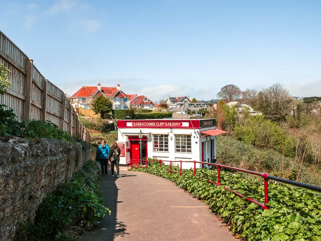 A wide path, with a stone wall and wooden fence on the left and metal railing and bushes to the right. The white and red Babbacombe Cliff Railway building is ahead with two people walking towards it.