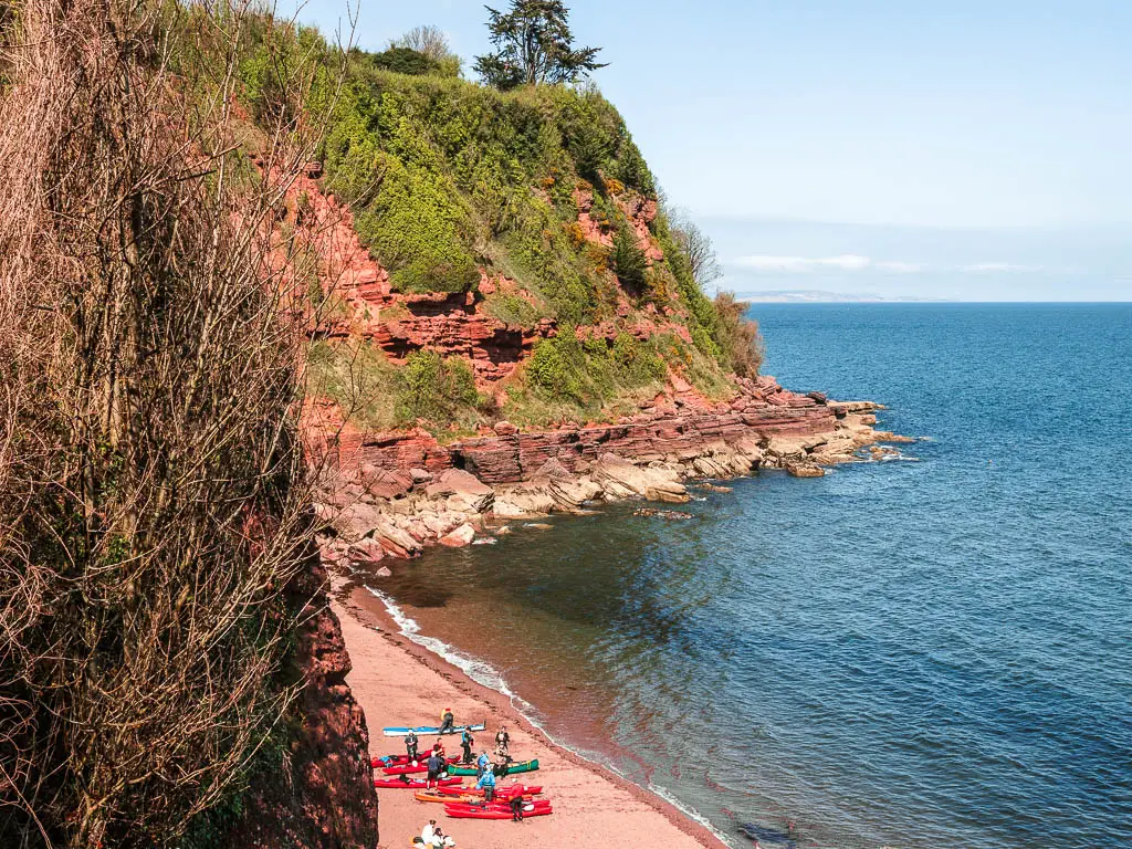 Looking down to the red sand beach of Maidencombe, completely surrounded by tall rugged cliffs on the coastal walk from Babbacombe to Teignmouth. There is a group of kayakers on the beach.