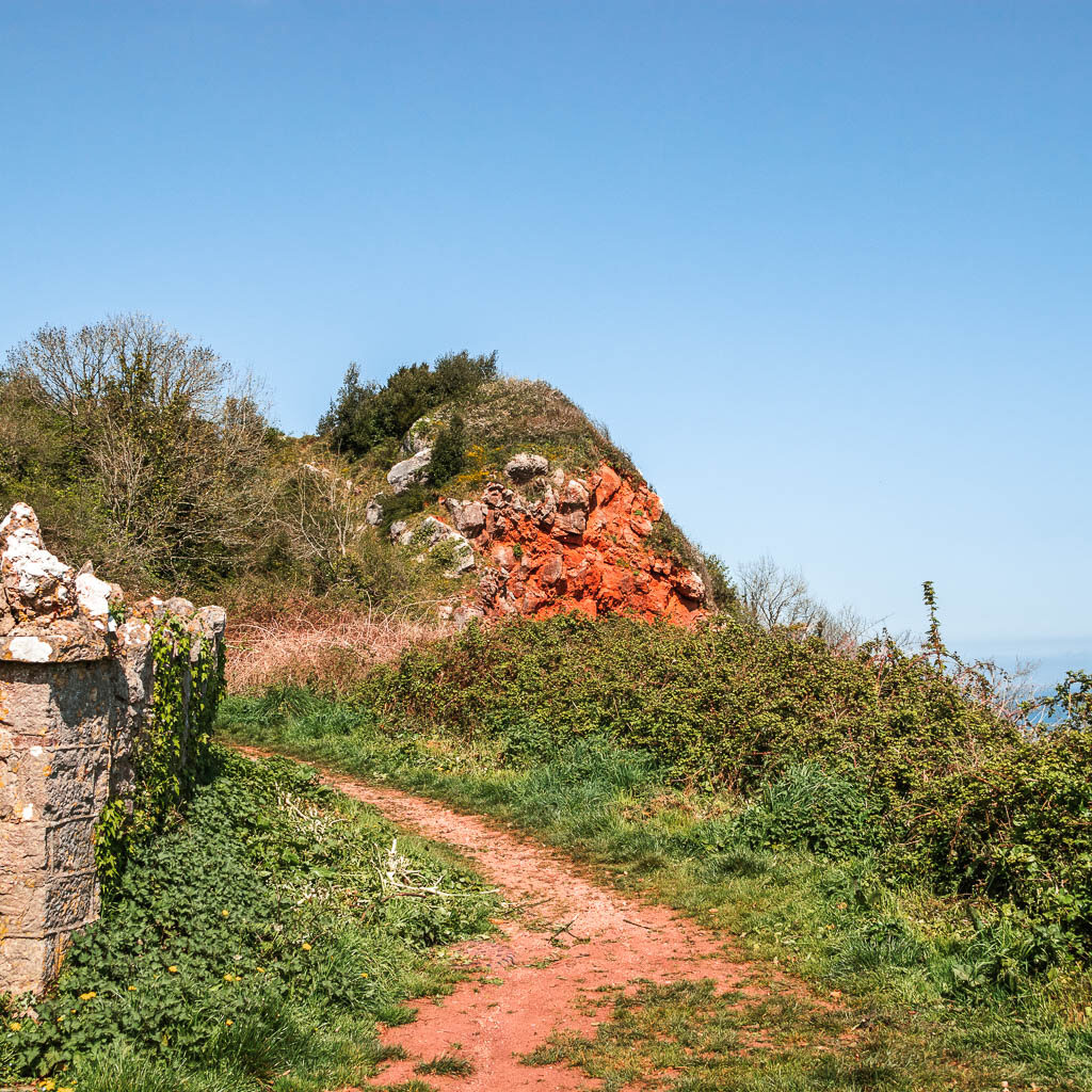 A dirt path through the grass and a bit of red cliff poking up ahead. 