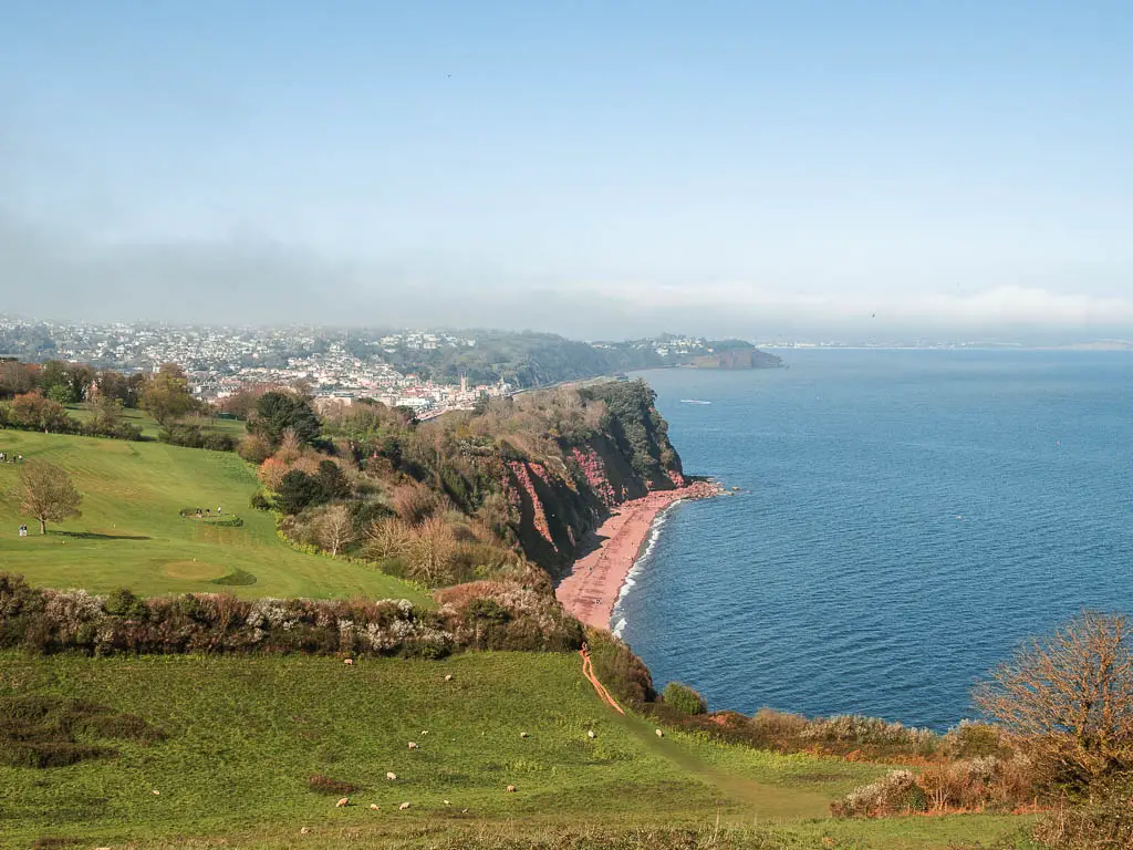 Looking down the green grass field on the clifftop with a view to Ness Beach, a long stretch of red sand, at the end of the coastal walk from Babbacombe to Teignmouth.