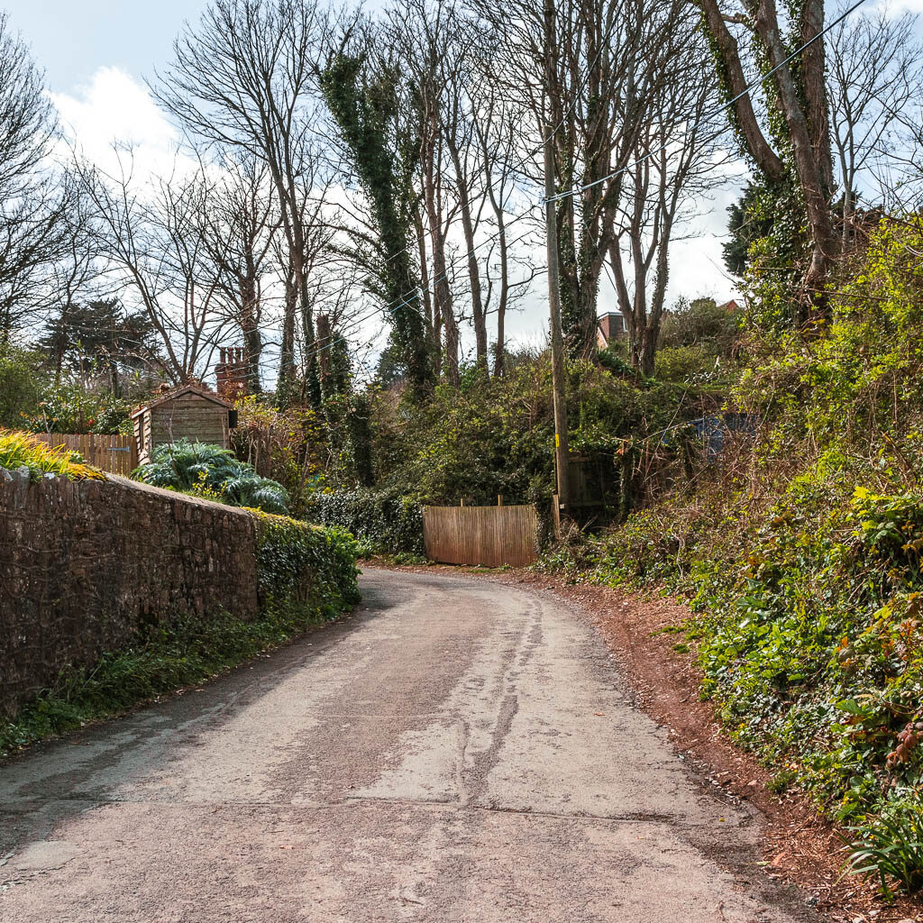 A road leading ahead and to the left. There is a stone wall on the left side and green bushes on the right.