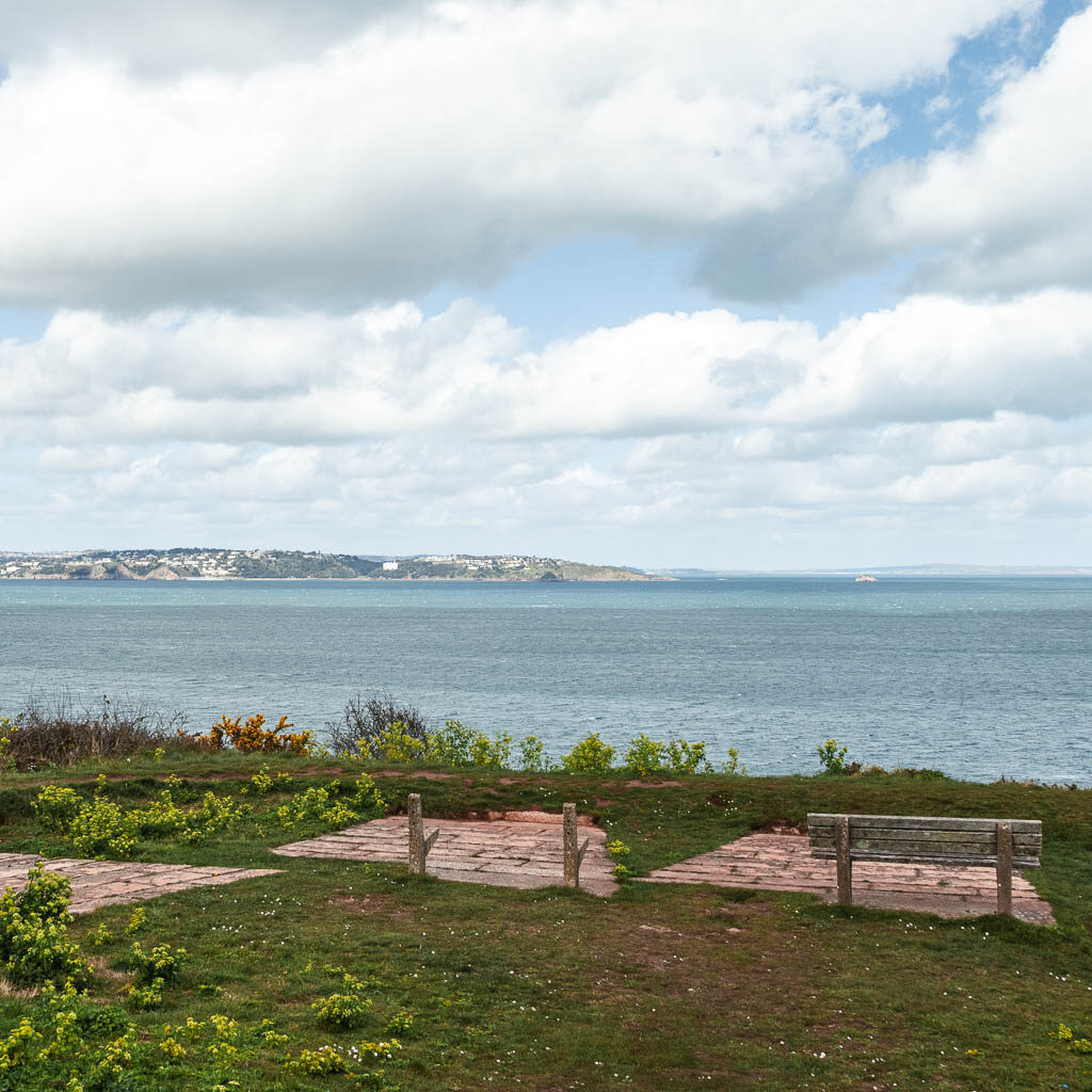 Looking across the green with a bench, towards the blue sea ahead and some land visible in the distance.