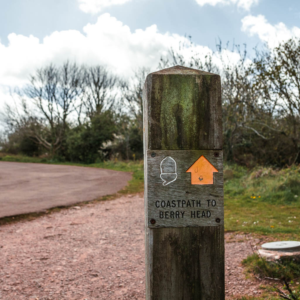 A wooden trial signpost which says 'coast path to Berry Head' along the walk from Brixham Harbour. The sign also has a yellow arrow and white acorn.