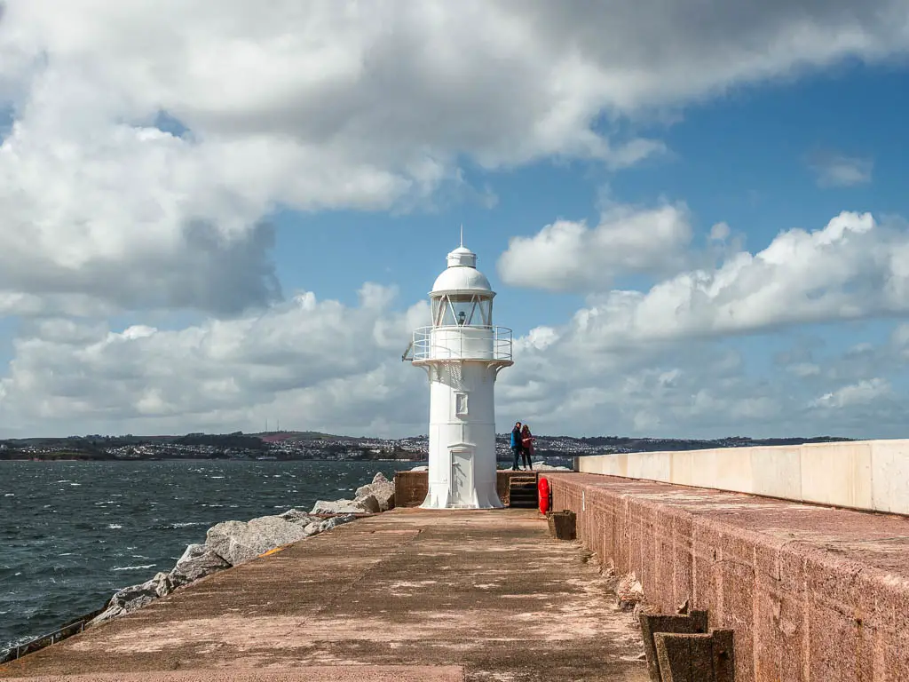 The white Brixham Lighthouse at the end of the walkway, with the sea to the and Brixham Harbour to the left.