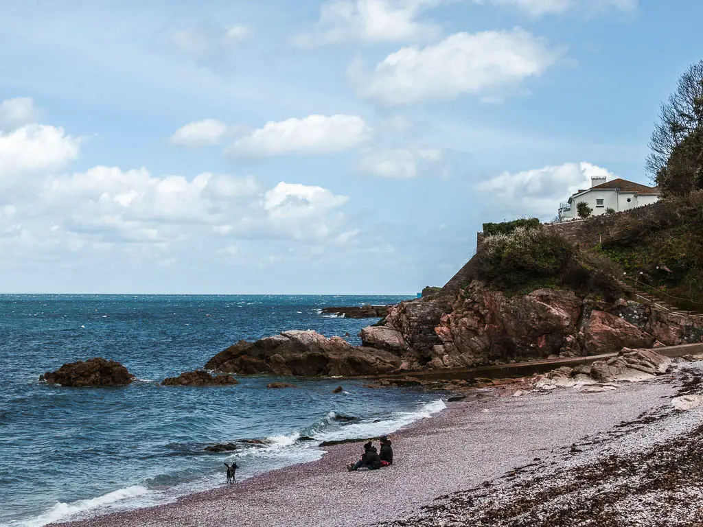 Looking down to the beach as it meets the blue sea, with rocks on the other side. There are two people sitting on the beach with their dog.