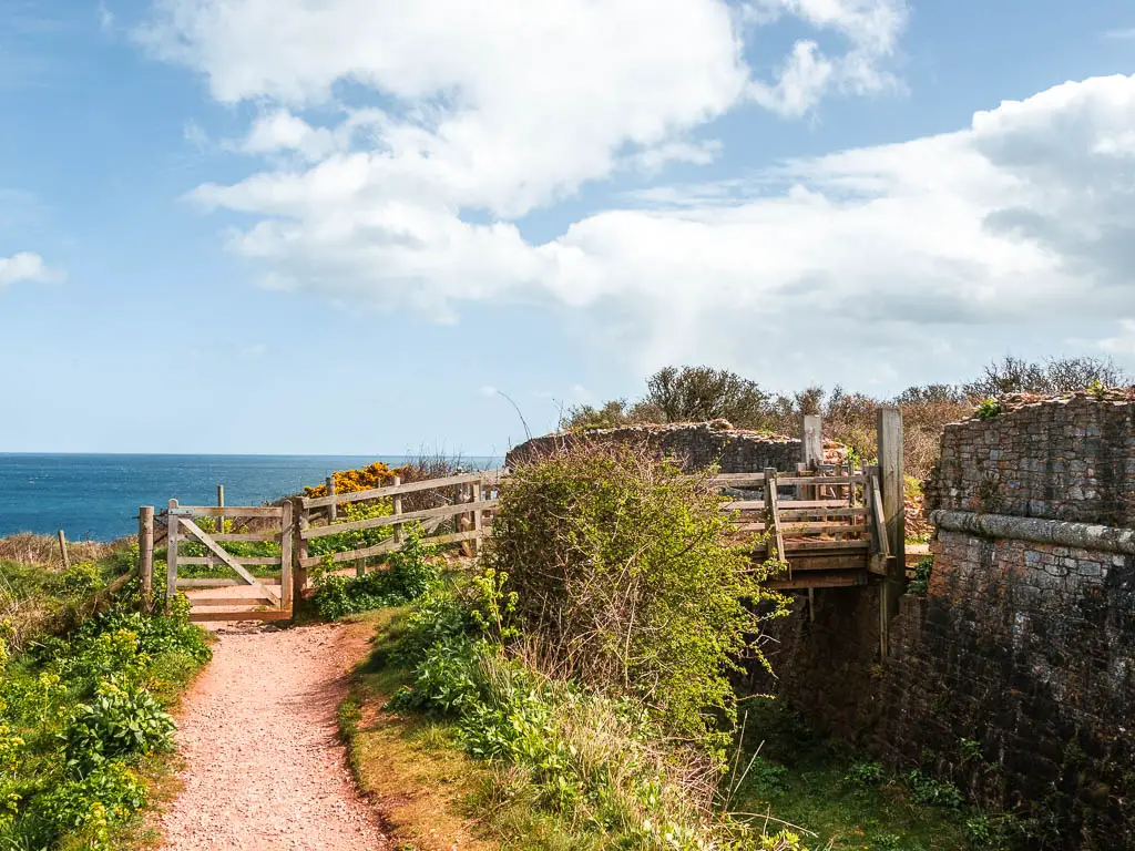 A gravel path on the left leading to a wooden fence and gate, with the ruins of a stone wall next to the ditch on the right.