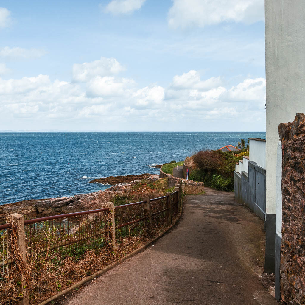 A path winding down with a metal fence on the left of it and the blue sea to the left and ahead.