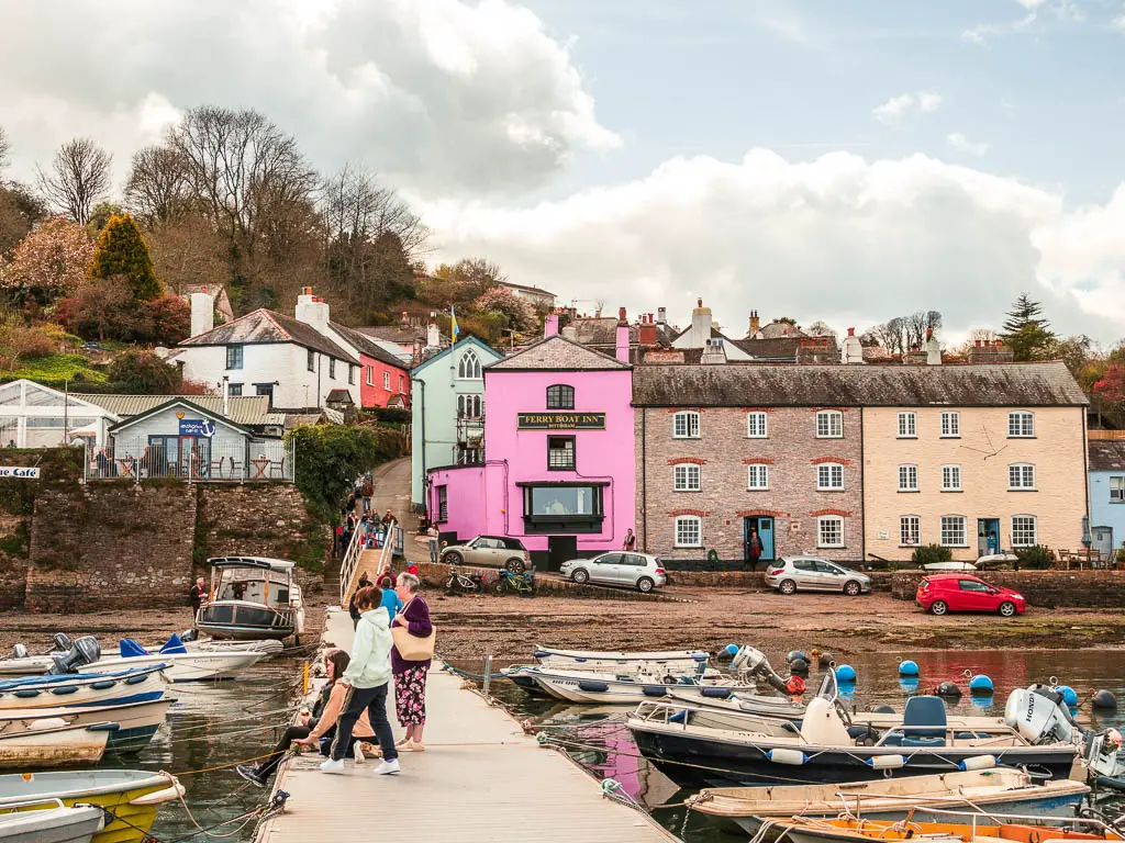 Looking along the pier towards the colourful facades of Dittisham partway through the walk from Dartmouth. There are a few people standing on the pier, and boats moored alongside it.